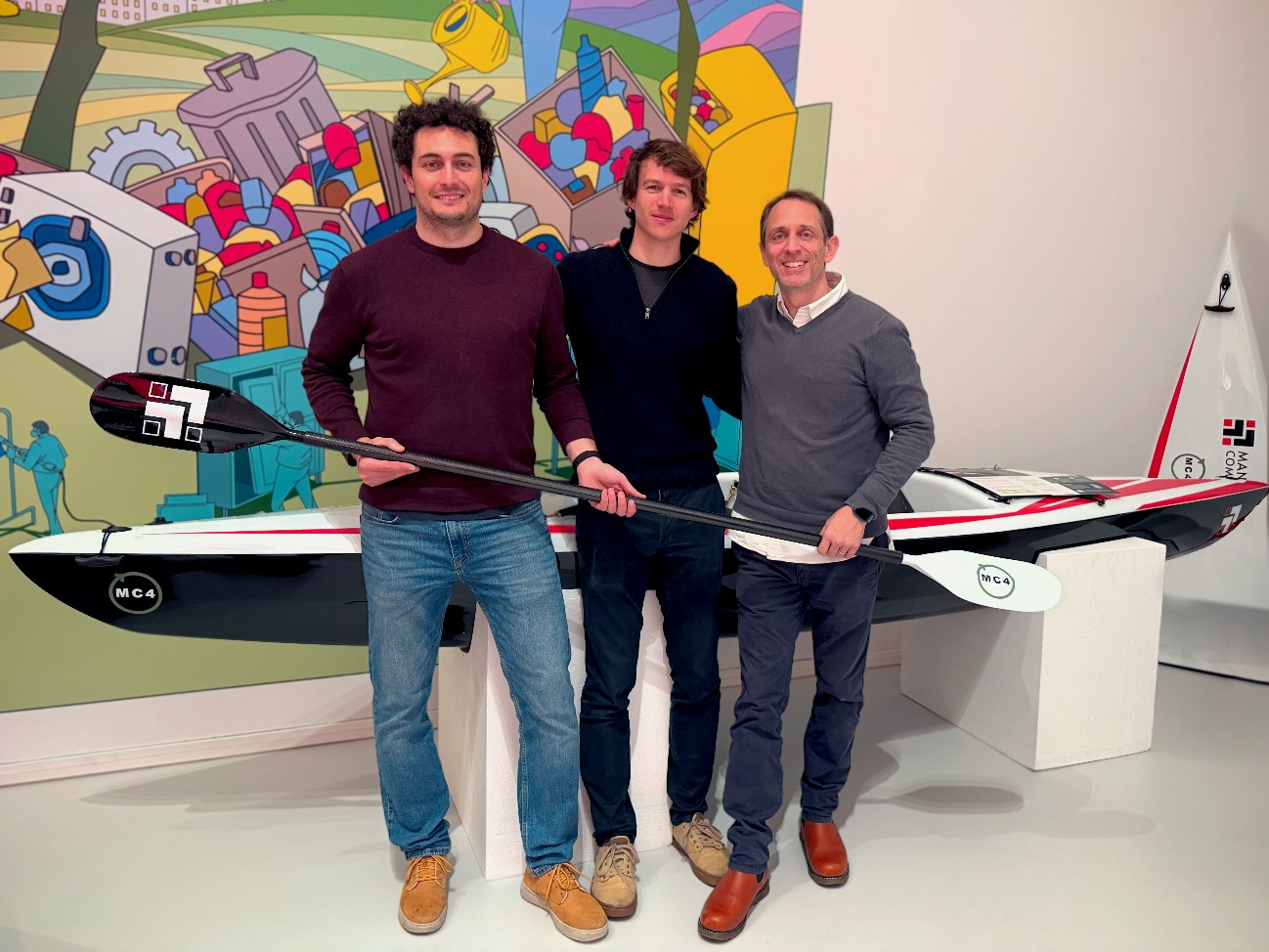 Asier Martín, Ronan Lecoeuche, and Eduardo Nicolás, the Managing Composites representatives in charge of the project presentatio, pose in front of the repurposed composite kayak during the final review presentation of the MC4 project. They are holding a kayak paddle, standing against a colorful illustrated backdrop with industrial and recycling themes