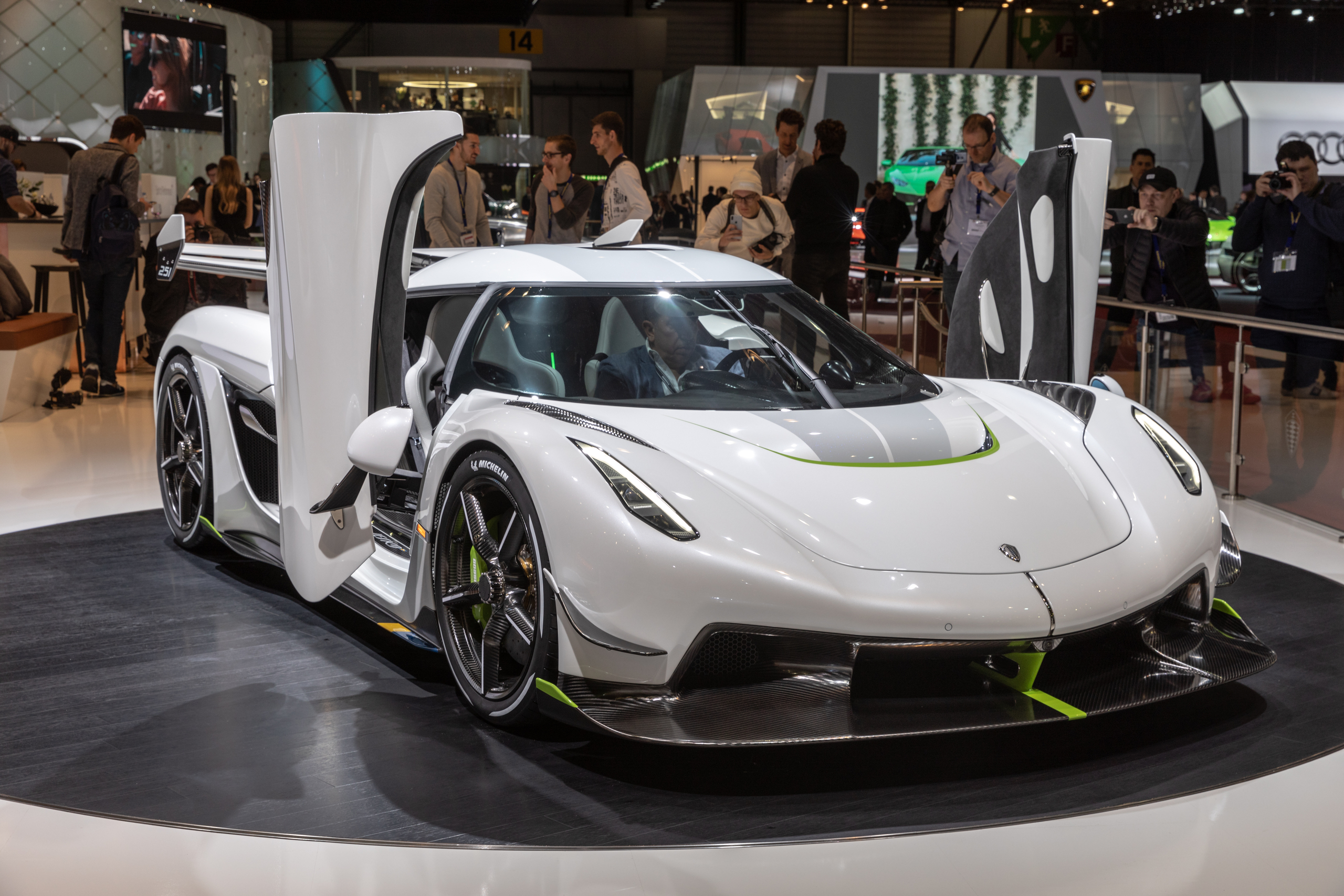 White Koenigsegg Jesko hypercar displayed at an auto show, with both dihedral doors open and several people observing and photographing the vehicle in the background