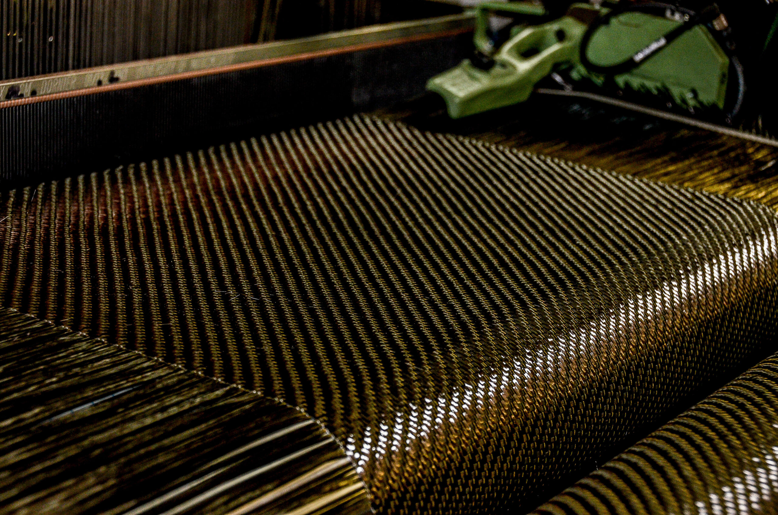 This photo shows a close-up view of carbon fiber being woven on an industrial loom. The tightly interlaced filaments create a distinctive, glossy pattern characteristic of carbon fiber fabrics.