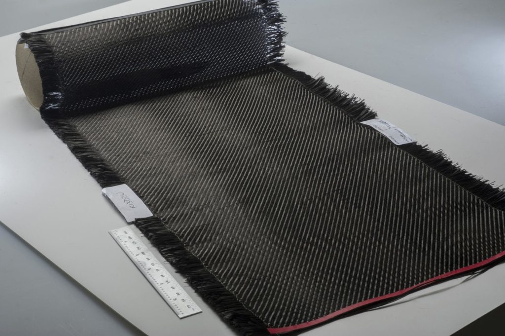 Roll of carbon-fiber fabric unrolled on a white surface, showing frayed edges and a diagonal weave pattern, with a ruler placed beside it for scale